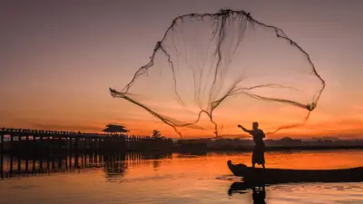 Silhouette of a man casting a large fishing net in the air from a boat during sunrise