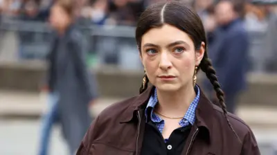 Lorde pictured in March. The singer has her dark hair braided in two plaits either side of her head. She wears gold shell-shaped earrings and a brown jacket over a checked blue shirt. She's pictured outside, with people walking behind her in the distance. 