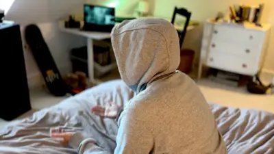 A young person wearing a grey hoodie is sitting on a bed in a student bedroom with computer screens on a desk in the background