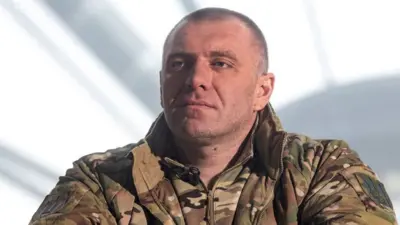 Чоловік середніх років у камуфляжній військовій формі спирається, схрестивши руки, на деревʼяну поверхню. На рукаві видно нашивку, а на зап’ясті – чорний годинник. Фон розмитий, але помітні великі металеві конструкції або ж вікна.