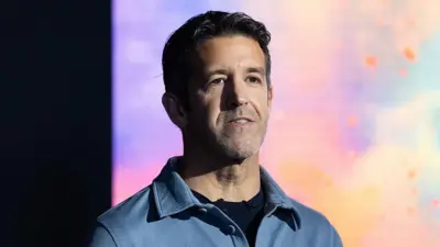 John Ternus veste uma camisa azul claro sobre uma camiseta azul marinho e está diante de um fundo colorido em tons de azul, roxo, amarelo, laranja e rosa