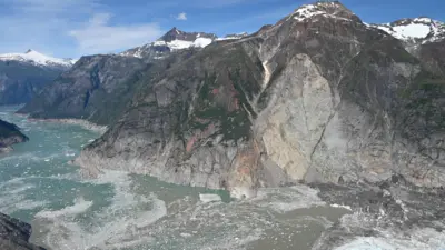 Dağlar arasındaki dar bir geçitten buzlu bir deniz uzanıyor. Dağların dik yamaçlarının büyük bölümü yeşil bitki örtüsünden arınmış; bunun yerine gri, parçalanmış kayalıklar ortaya çıkmış durumda. Dağların zirveleri karla kaplı ve aşağıdaki fiyortta buzlar parıldıyor.