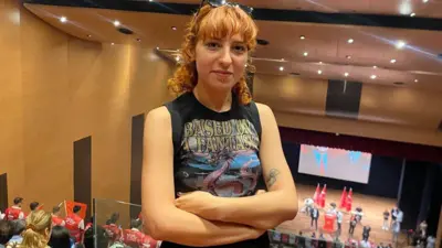Turuncu sarı saçlı genç bir kız, ellerini önünde kavuşturmuş fotoğraf çektiriyor. Kolsuz bir tişört giymiş ve saçlar kahkul. Arkada CHP bayrakları ve bir kürsü görünüyor.