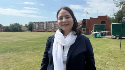 A woman with long dark brown hair wearing a blue jacket, a white scarf and a white top. She is standing on a grass sports field.