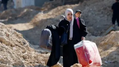 A woman carries a big bag in each hand walking between mounds of rubble on either side