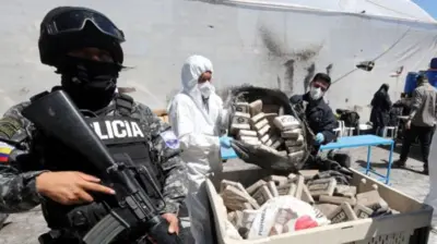 Police with guns stand guard over a container filled with drug packages as two people in masks and hazard clothes tip more drugs inside