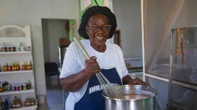 Novella Payne, wearing glasses and a hair covering, stirs a pot containing a sauce she makes using traditional recipes