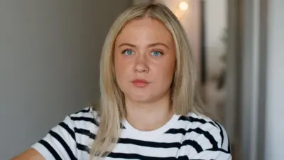Uma mulher loira com um piercing no nariz está olhando para a câmera. Ela está vestindo uma camiseta listrada preta e branca e está dentrobwin 216.netum prédio indefinido