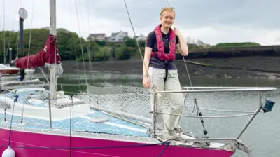 A young woman, wearing a pink life jacket with sun glasses on top of her head, stands on the bow of her pink boat looks at the camera