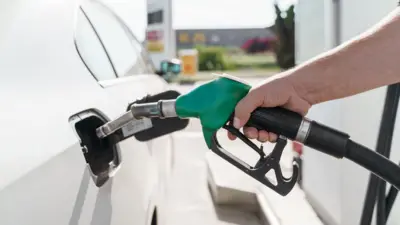 Рука людини тримає паливний пістолет із зеленою ручкою та заправляє білий автомобіль.