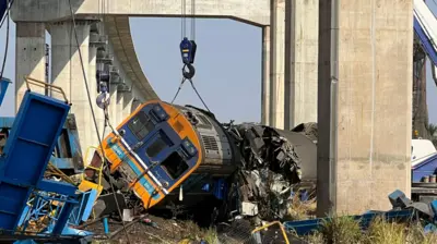 A damaged train lying on its side surrounded by twisted metal with overhead rail tracks in the background