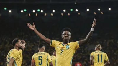 Vini Jr., com as mãos para o alto, acompanhado de outros jogadores da seleção brasileira de costas, todos uniformizados, em um estádio de futebol lotado