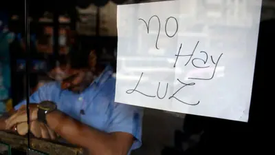 Un empleado de un negocio cerrado durante un apagón permanece detrás de la puerta con un letrero que dice: "No hay luz", en Caracas.