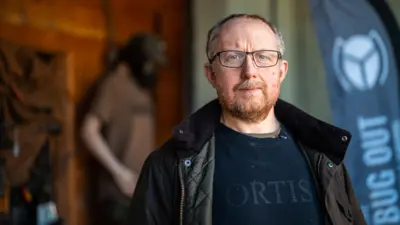 Leigh, um homem com barba ruiva e cabelo grisalho, está na varanda de sua oficina, vestindo uma jaqueta verde-escura sobre uma camisa preta