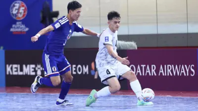 بازیکن افغانستان با لباس سفید پا به توپ، بازیکن جاپان با لباس نیلی و سفید به دنبال اوست.