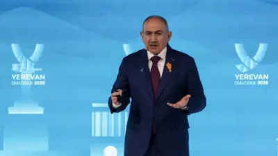 Nikol Paşinyan bir etkinlikte konuşuyor. Üzerinde takım elbise ve yakasında turuncu renkli Ermenistan haritası şeklinde rozet var.