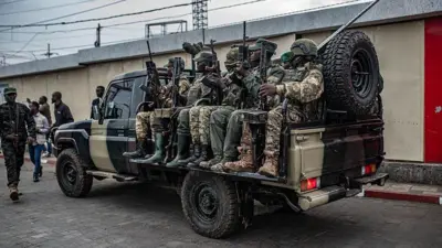 Abarwanyi bo mu mutwe w'inyeshyamba wa M23 bicaye mu modoka ya 'pickup', i Goma, ku itariki ya 6 Gashyantare (2) mu 2026. Bambaye imyenda yijimye, iruhande rwabo hari abandi bantu, n'umwe muri bo na we ufite imbunda arimo kugenda. Abicaye mu modoka bose bafite imbunda. Inyuma hari urukuta rw'inyubako yubakishije isima.