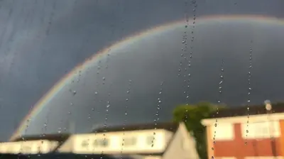 Looking through a window pane with raindrops running down it. Outside the window there are dark clouds and a rainbow