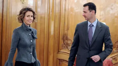 Asma al-Assad, de pelo corto y rizado, lleva una chaqueta gris y mira hacia un lado mientras su marido Bashar, con traje gris oscuro y corbata, la mira. Están de pie en una habitación revestida de madera con asientos con cojines rojos al fondo. Esta es una foto de archivo de 2010.