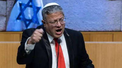 Itamar Ben-Gvir wearing a black suit, red tie and white kippah, talking on a podium in front of Israeli flags
