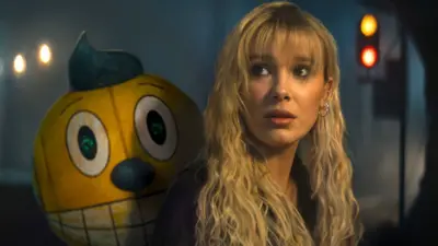 Millie Bobby Brown next to a robot companion with a yellow cartoon face with large eyes and mouth in The Electric State