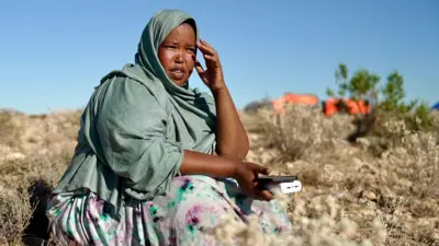 A woman sitting on a mountainside wearing a light grey scarf covering her head and shoulders. She is looking into the distance and holding a mobile phone on her lap.