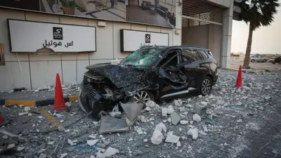 سيارة سوداء محطمة تقف بين مخروطين مروريين أحمرين وسط الأنقاض خارج فندق في المنامة، البحرين، يوم الأحد 1 مارس/آذار 2026، مع وجود سيارتين أخريين خلف شجرة نخيل في الخلفية. 