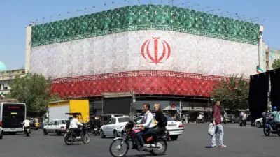 エンケラブ広場に臨む建物の二つの壁面にイラン国旗が大きく掲示されている。その前を車両や歩行者が行きかう