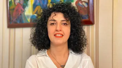 Narges Mohammadi sonríe en esta foto de folleto sin fecha. Tiene el pelo negro rizado y lleva una camisa blanca, un collar en forma de triángulo y lápiz labial rojo.