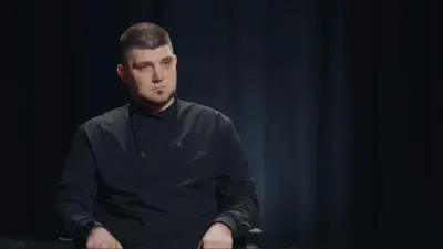 Капустін сидить у студійному кріслі на темному фоні. Він одягнений у темну сорочку, має коротку стрижку та бороду. Вираз обличчя серйозний і зосереджений, освітлення м’яке й спрямоване на нього.
