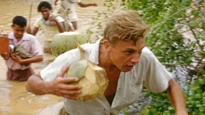 An image of a young Attenborough crossing a body of water. Three men can be seen in the background behind him.