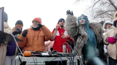 Женщины и мужчины в яркой теплой одежде танцуют вокруг диджея на морозе