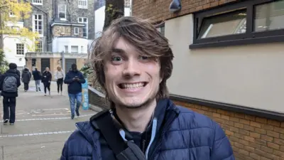 A smiling Gillie Owen with brown hair, wearing a blue puffa jacket and shoulder strap bag