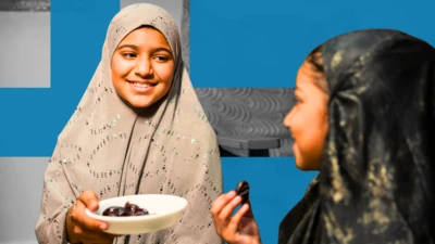 دوې نجونې چې په حجاب کې دي، د یو بل تر څنګ موسکی ناستې دي. د یوې په لاس کې له خورما ډک کاب دی.