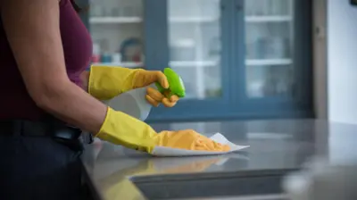 Mulher de luva joga produto com spray em mesa; não é possível ver seu rosto