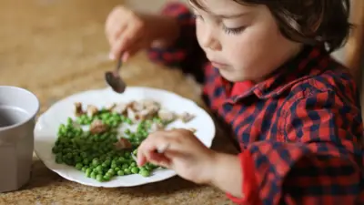 A child with brown hair in a red and black chequered shirt sits at a table and eats a plate of peas and mushrooms