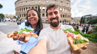 Egy férfi és egy nő pizza szeleteket tart a kezében a római Colosseum előtt.