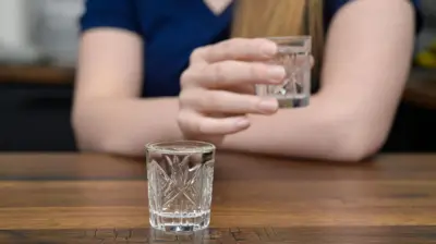 glass containing clear liquid on a table with a woman holding a similar glass in the background