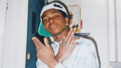 Mark Wilberforce smiles at the camera as a teenager in London wearing a white cap backwards, a gold earring and a white checked shirt. His hands are crossed and held up with the last two fingers on each bent.