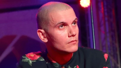 Николай Комягин в черной рубашке с красными цветами, наголо обритый, смотрит куда-то вверх. Задний план сиреневого цвета 