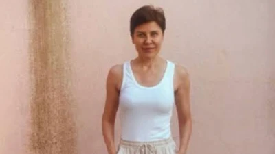 Beyaz atlet giyen bir kadın, turuncu bir duvarın önünde elleri eşfomanının cebinde duruyor. Saçları çok kısa kesilmiş durumda.