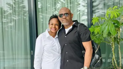 Tony Elumelu wife Awele wear white shirt while Tony wer black shirt wit black eyeglass and carry black bag. Di wife lean for im body.
