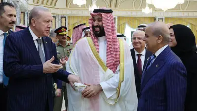Cumhurbaşkanı Erdoğan solda, Muhammed bin Salman ortada, Şahbaz Şerif sağda. Üç lider yürürken gülüşerek sohbet ediyor. Etraflarında danışmanları ve askeri üniformalı kişiler var. Erdoğan ve Şerif takım elbise giyiyor, bin Salman'ın üzerinde geleneksel Suudi Arabistan kıyafeti var. Arka planda sarı renkli duvarlar görünüyor.