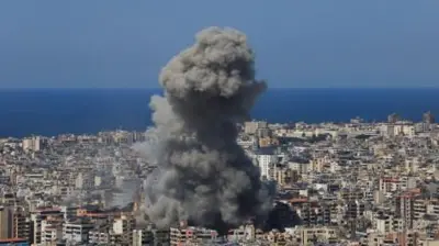 日中の市街地で、大きな黒煙が上っている。建物が密集している。背景に海が見える