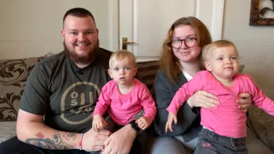 A dad with a beard, wearing a dark t-shirt, sits on a sofa alongside his wife, who has long ginger hair and wears a grey cardigan. Their twin daughters, wearing matching pink long-sleeved t-shirts, sit on their knees.