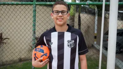 Um menino usando óculos de grau e uma camisa de futebol do Ceará. Ele segura uma bola de futebol com a mão direita.