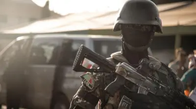 Um militar do Mali usando capacete, máscara facial e óculos escuros olhando para a câmera e segurando um rifle