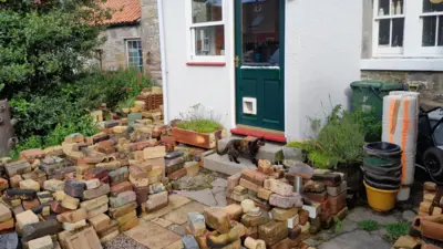 Ian Suddaby's garden is covered in piles of colourful bricks. There is a cat on his doorstep and his door is green and has a glass pane in it,