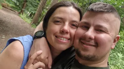 Josh, pria berambut cokelat dengan janggut tipis mengenakan kaus hijau, berfoto selfie dengan merangkul leher seorang perempuan berambut cokelat, Kelly mengenakan atasan tanpa lengan biru muda dan meletakkan tangannya di atas tangan Josh. Mereka berdiri di hutan dengan pepohonan di latar belakang, keduanya tersenyum dan menatap kamera.