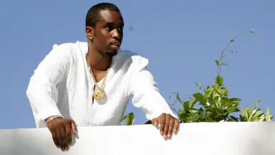 Sean "Diddy" Combs looks out over a white balcony wall while wearing a white top and gold chain, with a plant next to him and blue sky in the background, at one of his famous White Parties at his East Hapton estate in New York in 2007